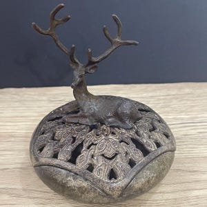 Puede incluir: Un quemador de incienso decorativo de color bronce con una detallada figura de ciervo sobre una base esférica calada. La base tiene un intrincado patrón simétrico. El ciervo tiene astas prominentes y está en posición de descanso.