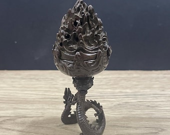 May include: A dark brown incense burner with intricate carvings in the shape of a dragon. The burner is made of metal and has a round, open top with a decorative design. The dragon is curled around the base of the burner, with its head facing upwards.