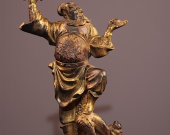 Op de afbeelding: Bronzen beeld van een krijger in een dynamische pose, met een zwaard omhoog. De figuur draagt een gedetailleerd harnas. Een kleiner figuur knielt aan de voeten van de krijger. Het beeld heeft een goud- en bronskleur.