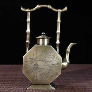 May include: A brass teapot with a bamboo handle and a decorative engraved design of a mountain landscape and a pagoda. The teapot has an octagonal body and a lid with a knob.