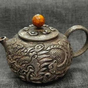 Può includere: Teiera d'argento con intricate incisioni a rilievo di draghi e nuvole. Il coperchio presenta un piccolo pomello rotondo marrone-arancio. Il manico e il beccuccio sono argentati. Un oggetto decorativo.