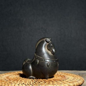 Puede incluir: Figura de caballo gris oscuro con cuerpo redondeado y melena y rostro detallados. El caballo lleva un arnés decorativo y está sentado sobre una estera circular tejida. El fondo es de color oscuro.