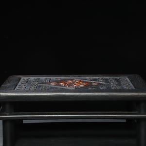 May include: A dark grey wooden table with a rectangular top and four short legs. The top features carved Chinese characters and a central design in reddish-brown. The table is a low, decorative piece.