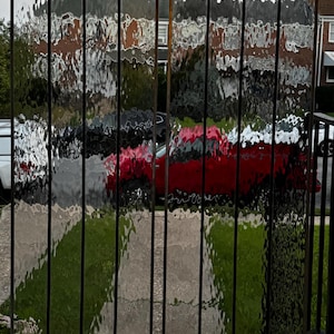 Puede incluir: Un par de puertas de vidrio con marco de metal negro con un diseño de vidrio texturizado. El vidrio es transparente y tiene un patrón ondulado. Las puertas están cerradas y se puede ver un coche rojo y hierba verde a través del vidrio.