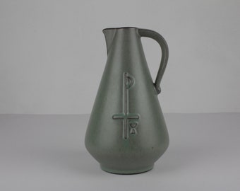 May include: A green ceramic pitcher with a stylized design on the front. The design features a cross with a circle at the top and a small triangle at the bottom.