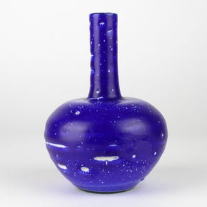 Puede incluir: Un jarrón de cerámica azul cobalto con un cuerpo redondeado y un cuello largo y estrecho. El jarrón tiene un patrón moteado de puntos blancos y vetas blancas irregulares. El cuello tiene un diseño texturizado y acanalado.