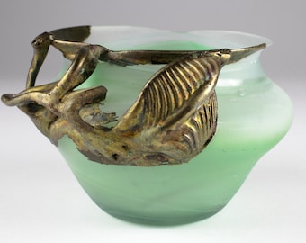 May include: A small, green glass vase with a decorative, gold-toned metal handle. The handle is shaped like a stylised leaf and has a textured surface.
