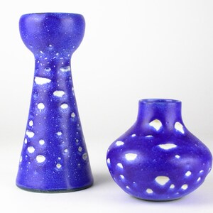 Op de afbeelding: Twee blauwe keramische vazen met witte stippen. De hogere vaas heeft een uitlopende bovenkant en een smalle basis. De kortere vaas is ronder met een bredere basis.