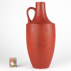 Puede incluir: Un jarrón de cerámica rojo alto y texturizado, con una sola asa. El jarrón mide aproximadamente 30 cm de alto.