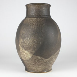 Pode incluir: Um vaso de cerâmica castanho escuro com corpo arredondado e abertura ligeiramente alargada. O vaso tem uma superfície texturizada salpicada com toques de castanho claro e branco.