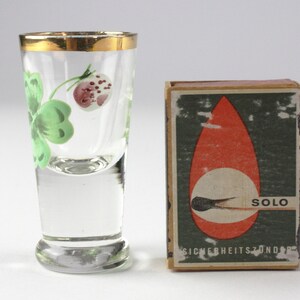May include: A clear shot glass with a gold rim and a painted design of a green clover and a red berry. Next to it is a matchbox with the word "SOLO" and the text "SICHERHEITSZONDER" printed on it.