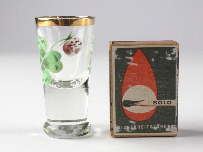 May include: A clear shot glass with a gold rim and a painted design of a green clover and a red berry. Next to it is a matchbox with the word "SOLO" and the text "SICHERHEITSZONDER" printed on it.