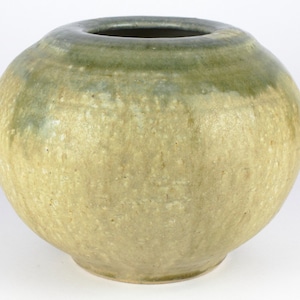 Può includere: Un vaso rotondo in ceramica con una smaltatura verde chiaro. La smaltatura ha un effetto screziato, con aree di verde più scuro e giallo più chiaro. Il vaso ha un'apertura larga e un fondo arrotondato.