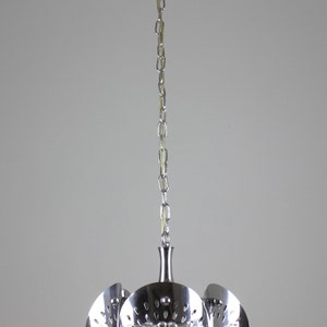May include: Chrome pendant light fixture with a decorative floral design. The light has a series of curved, perforated metal shades and a central glass element. The fixture hangs from a chain.