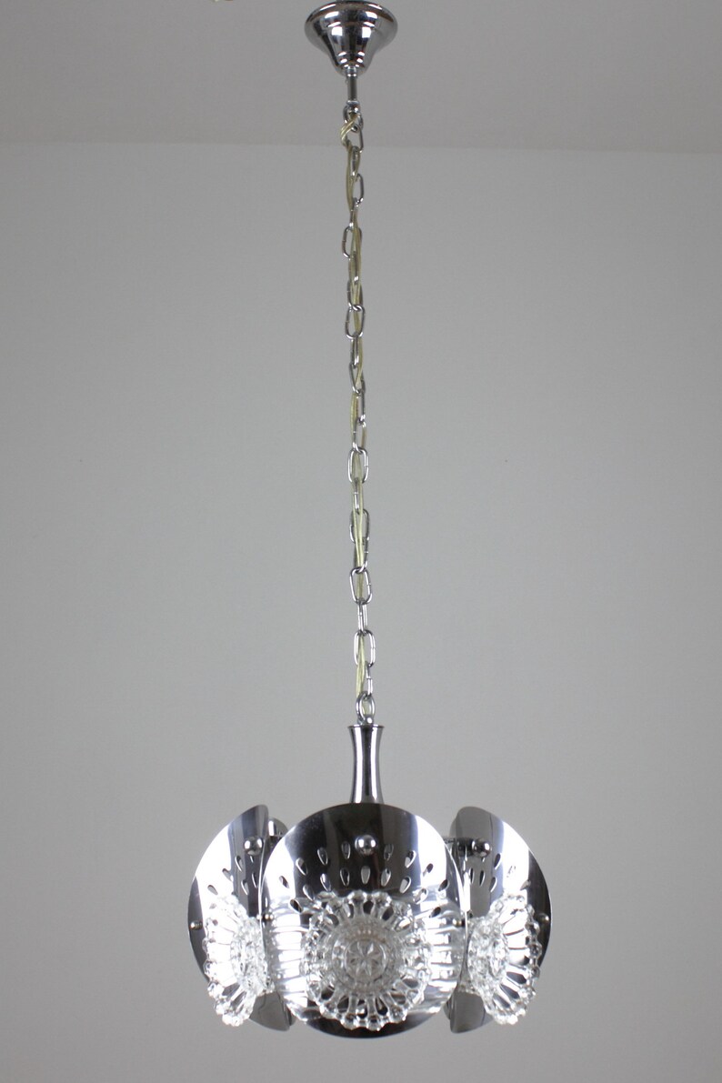 May include: Chrome pendant light fixture with a decorative floral design. The light has a series of curved, perforated metal shades and a central glass element. The fixture hangs from a chain.