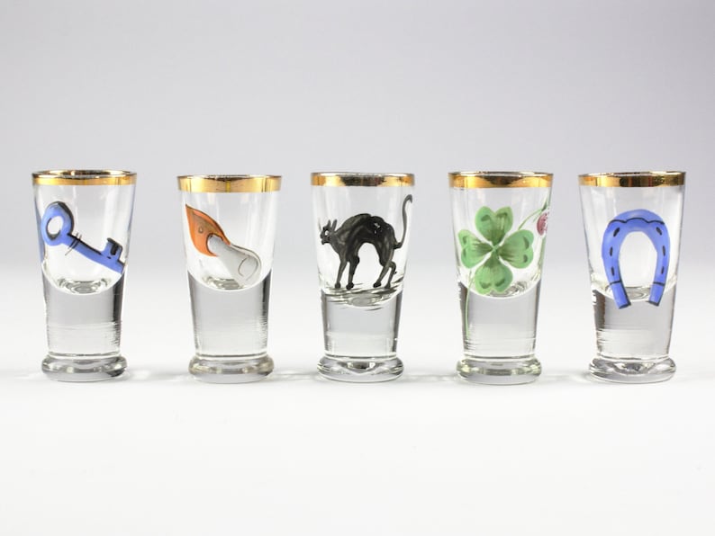 May include: Five clear shot glasses with gold rims, each featuring a different hand-painted design: a blue key, an orange and white object, a black cat, a green four-leaf clover, and a blue horseshoe. The glasses are arranged in a row.