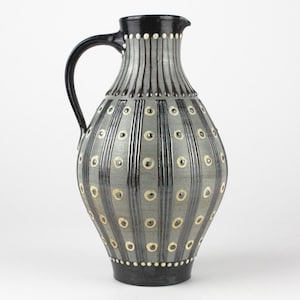 May include: A tall, ceramic pitcher with a black handle and rim. The body is gray with vertical black stripes and small, evenly spaced, circular cream-colored accents. The base is black, and the pitcher has a spout for pouring.