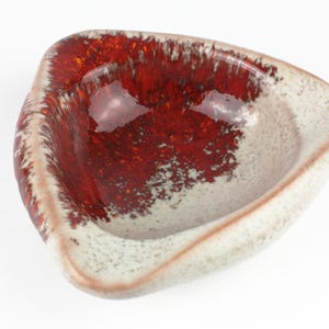 Può includere: Una ciotola triangolare in ceramica con esterno screziato bianco sporco e interno rosso intenso lucido. Il bordo ha un bordo marrone-rossastro. La ciotola è probabilmente per scopi decorativi o di servizio.