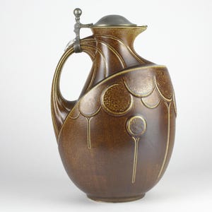 Op de afbeelding: Bruine keramische kan met een decoratief ontwerp. De kan heeft een handvat, een deksel en een tuit. Het ontwerp bevat cirkelvormige en verticale elementen. De kan is ongeveer 30 cm hoog.