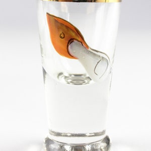 May include: Clear shot glass with a gold rim and a painted orange and white design. The design appears to be a stylized bird or swan. The glass is on a white background.