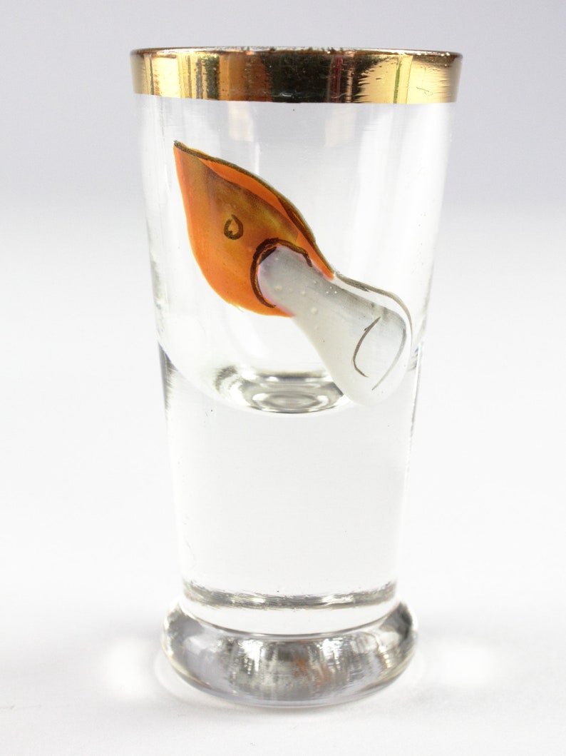 May include: Clear shot glass with a gold rim and a painted orange and white design. The design appears to be a stylized bird or swan. The glass is on a white background.