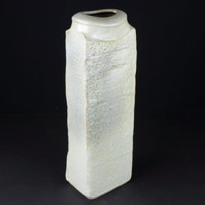 May include: A tall, rectangular vase with a textured, off-white surface. The vase has a slightly curved top with a small opening. The overall shape is geometric, and the texture gives it a rustic appearance.