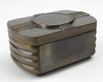 Pode incluir: Uma pequena caixa retangular de cor bronze com uma tampa decorativa. A tampa tem um design circular com um centro elevado e uma borda texturizada. A caixa tem um design nervurado nas laterais e assenta em quatro pequenos pés.