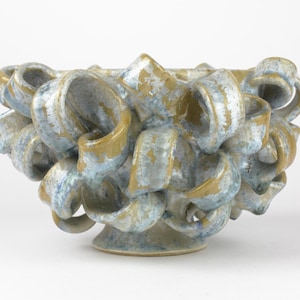 May include: A ceramic bowl with a unique, textured design. The bowl features a series of looped, ribbon-like forms in shades of blue, tan, and gold. The bowl sits on a small, circular base, and the overall shape is organic and sculptural.