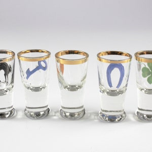 May include: Five clear shot glasses with gold rims, each featuring a different design: a black cat, a blue key, a horseshoe, and a green four-leaf clover. The glasses are arranged in a row.