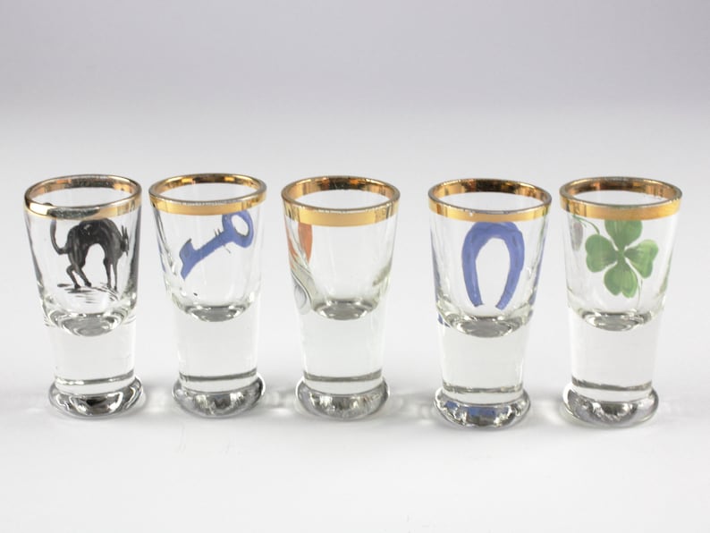 May include: Five clear shot glasses with gold rims, each featuring a different design: a black cat, a blue key, a horseshoe, and a green four-leaf clover. The glasses are arranged in a row.