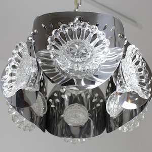 May include: A chrome-finished pendant light fixture with clear, faceted glass accents. The light has a layered design with multiple tiers of curved metal and decorative glass elements. The fixture is suspended from a chain and is suitable for interior lighting.