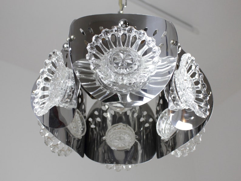May include: A chrome-finished pendant light fixture with clear, faceted glass accents. The light has a layered design with multiple tiers of curved metal and decorative glass elements. The fixture is suspended from a chain and is suitable for interior lighting.