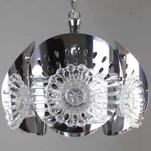 May include: A chrome-finished pendant light fixture with a unique design. The fixture features a central circular element with decorative cutouts, surrounded by curved, petal-like panels. Clear, ornate glass elements are incorporated for added detail.