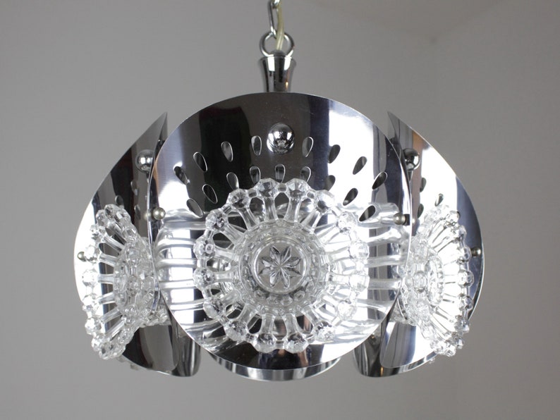 May include: A chrome-finished pendant light fixture with a unique design. The fixture features a central circular element with decorative cutouts, surrounded by curved, petal-like panels. Clear, ornate glass elements are incorporated for added detail.
