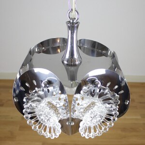 May include: A chrome-finished, mid-century modern pendant light fixture. The light has a central stem and a series of curved, perforated metal panels. Clear, faceted glass elements are suspended below the metal panels, adding a decorative touch.
