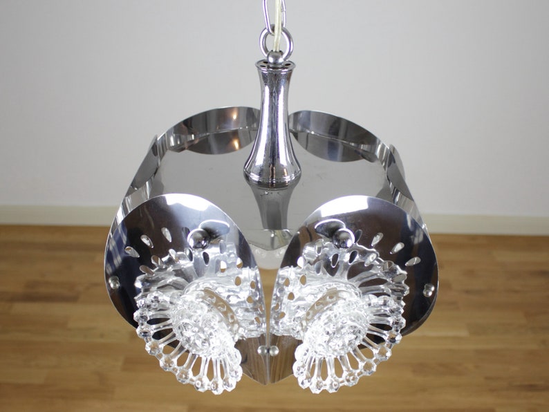 May include: A chrome-finished, mid-century modern pendant light fixture. The light has a central stem and a series of curved, perforated metal panels. Clear, faceted glass elements are suspended below the metal panels, adding a decorative touch.