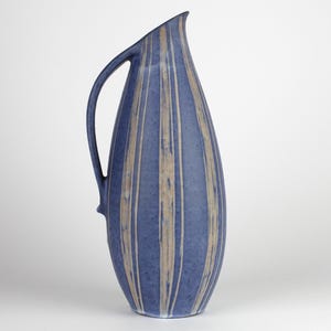 Puede incluir: Jarra de cerámica azul con asa y forma alargada. Rayas beige verticales decoran la jarra. Presenta un pico curvo y un pequeño elemento decorativo cerca de la base del asa. Mide aproximadamente 30 cm de altura.