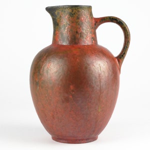 Puede incluir: Una jarra de cerámica roja y verde con una base ancha y un asa. La jarra tiene un esmalte moteado.