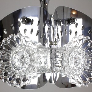 May include: A chrome and crystal chandelier with a unique design. The fixture features two large, curved, polished metal elements with oval cutouts, and sparkling crystal accents. The central crystal elements have a starburst pattern.
