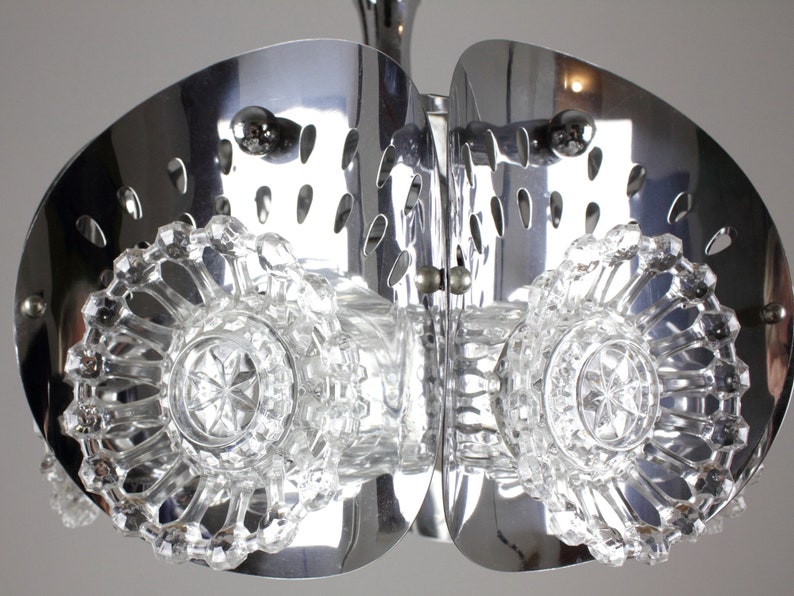 May include: A chrome and crystal chandelier with a unique design. The fixture features two large, curved, polished metal elements with oval cutouts, and sparkling crystal accents. The central crystal elements have a starburst pattern.