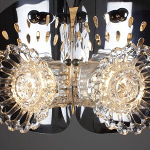 May include: A close-up of a modern chandelier with a polished chrome finish. The fixture features clear, faceted crystal accents and decorative cutouts. The design incorporates multiple light sources for a bright, elegant aesthetic.