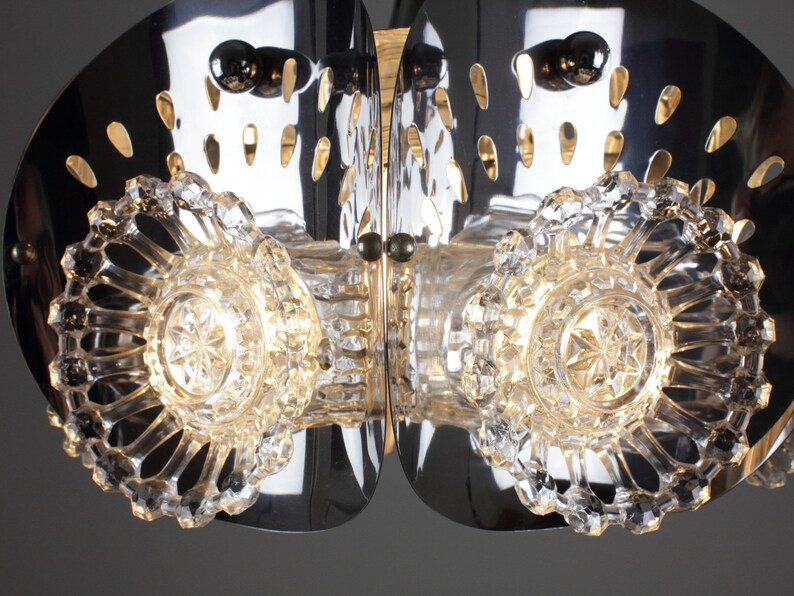 May include: A close-up of a modern chandelier with a polished chrome finish. The fixture features clear, faceted crystal accents and decorative cutouts. The design incorporates multiple light sources for a bright, elegant aesthetic.