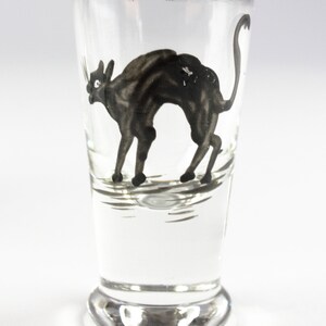 May include: Clear shot glass with a gold rim, featuring a black cat illustration. The cat is depicted in a stylized, arched pose. The glass has a rounded base and a straight, cylindrical body. The cat illustration is detailed with visible whiskers and a curved tail.