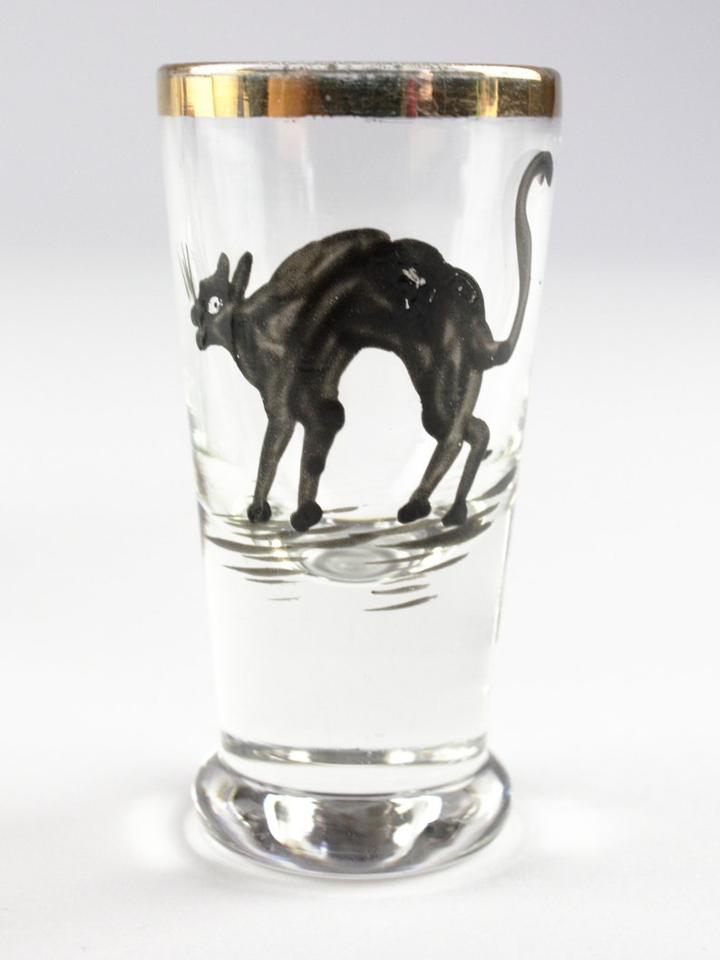 May include: Clear shot glass with a gold rim, featuring a black cat illustration. The cat is depicted in a stylized, arched pose. The glass has a rounded base and a straight, cylindrical body. The cat illustration is detailed with visible whiskers and a curved tail.