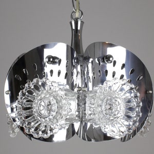 May include: A chrome-finished, mid-century modern chandelier with a unique design. The fixture features two large, semi-circular metal shades with decorative cutouts. Clear, faceted glass elements are suspended below, adding a touch of sparkle. The light fixture is suspended from a chrome chain.