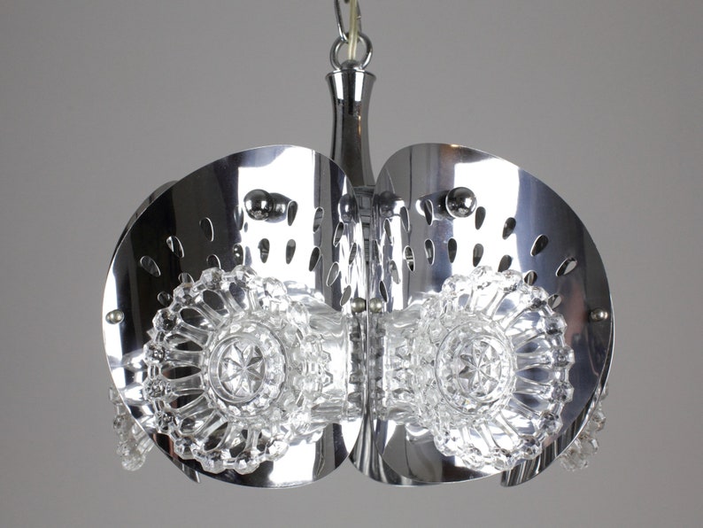 May include: A chrome-finished, mid-century modern chandelier with a unique design. The fixture features two large, semi-circular metal shades with decorative cutouts. Clear, faceted glass elements are suspended below, adding a touch of sparkle. The light fixture is suspended from a chrome chain.