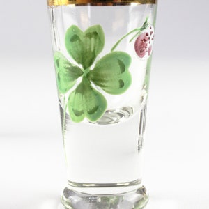 May include: A clear shot glass with a gold rim, featuring a hand-painted green four-leaf clover and a red berry. The glass has a flared base and is ideal for serving small portions of spirits or liqueurs.