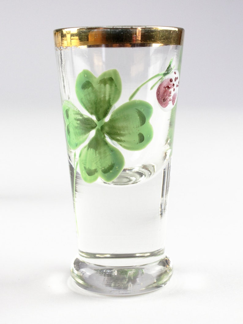 May include: A clear shot glass with a gold rim, featuring a hand-painted green four-leaf clover and a red berry. The glass has a flared base and is ideal for serving small portions of spirits or liqueurs.