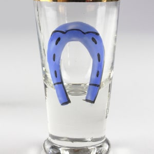 May include: A clear shot glass with a gold rim and a blue horseshoe design. The horseshoe is painted in a vibrant blue color with black accents. The glass is designed to hold a small amount of liquid.