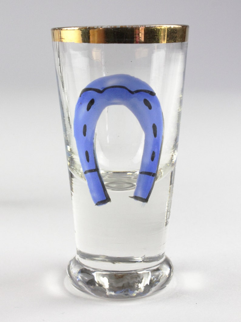 May include: A clear shot glass with a gold rim and a blue horseshoe design. The horseshoe is painted in a vibrant blue color with black accents. The glass is designed to hold a small amount of liquid.